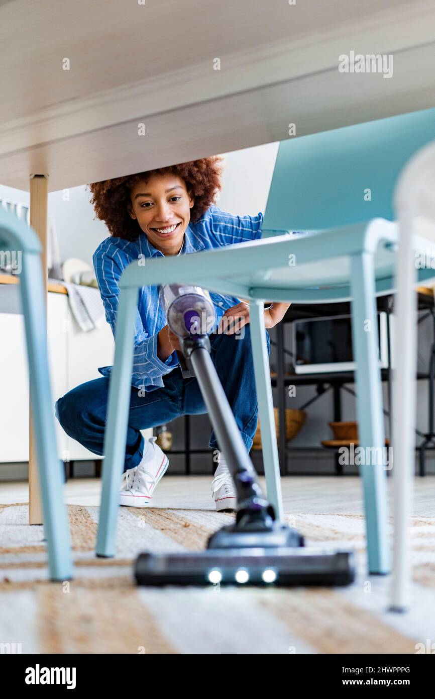 Une femme souriante se caroupe à l'aide d'un aspirateur sous une table à manger à la maison Banque D'Images