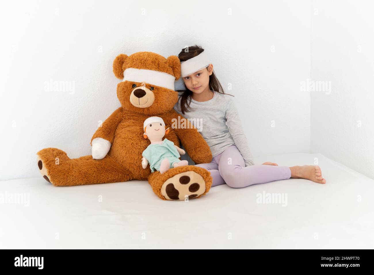 Bandage sur une fille avec un ours en peluche et une poupée assise devant le mur blanc Banque D'Images