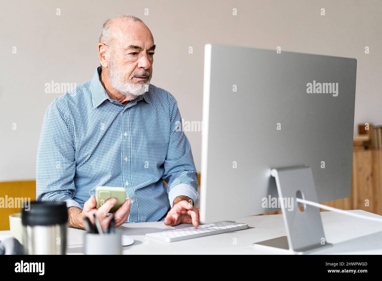 Indépendant senior tenant un smartphone travaillant sur un ordinateur au bureau à domicile Banque D'Images