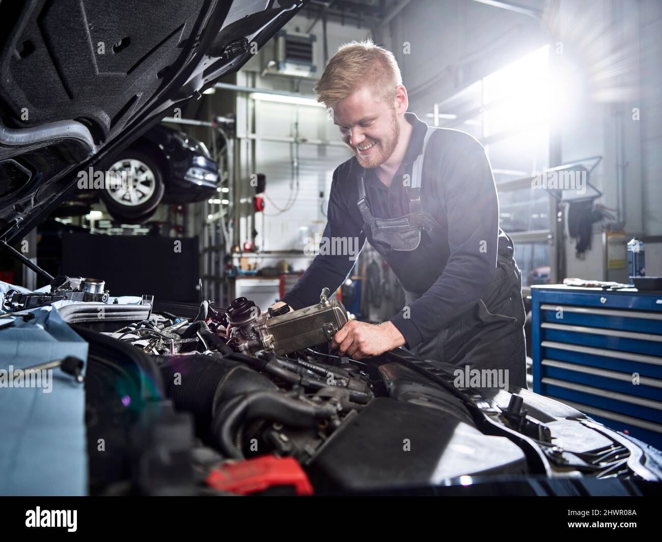 Un mécanicien blond souriant répare le moteur de la voiture dans un atelier automobile Banque D'Images