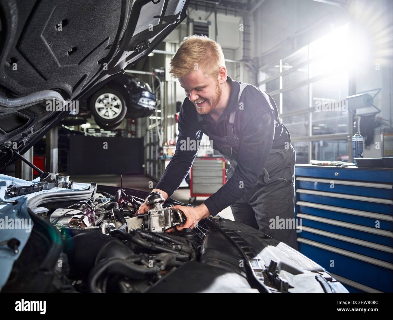 Jeune homme blond souriant mécanicien répare le moteur de voiture dans l'atelier automobile Banque D'Images