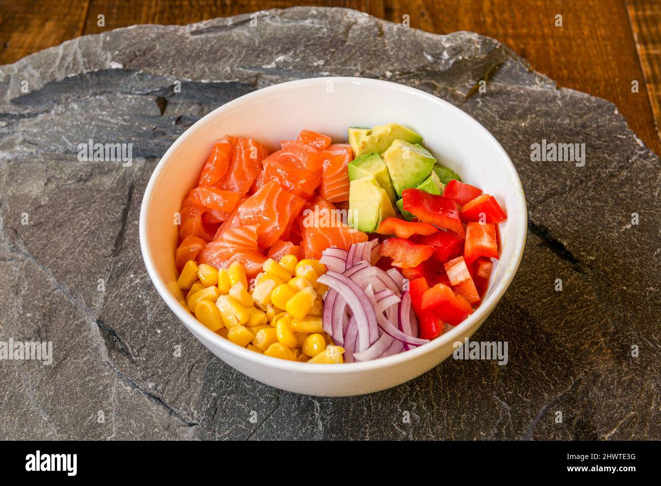 Bol à poke blanc avec cubes de saumon norvégien, maïs doux, morceaux de poivron rouge, dés d'avocat mexicain, riz blanc, oignon violet sur roche grise Banque D'Images