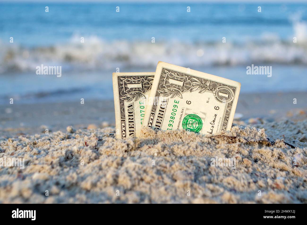 Deux billets d'un dollar à moitié ensevelis dans le sable sur le sable de bord de mer de gros plan. Banque D'Images