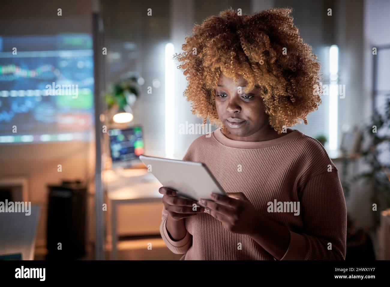 Jeune femme africaine aux cheveux bouclés regardant quelque chose sur une tablette numérique tout en travaillant au bureau jusqu'à tard dans la soirée Banque D'Images