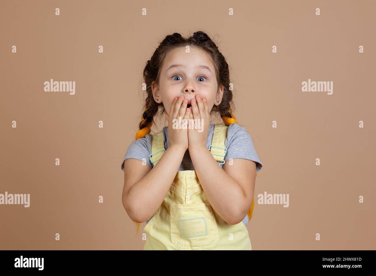 Portrait de la petite fille excitée choquée avec des tresses kanekalon, la fermeture de la bouche avec les mains faisant grimace regardant l'appareil photo portant la combinaison jaune et Banque D'Images