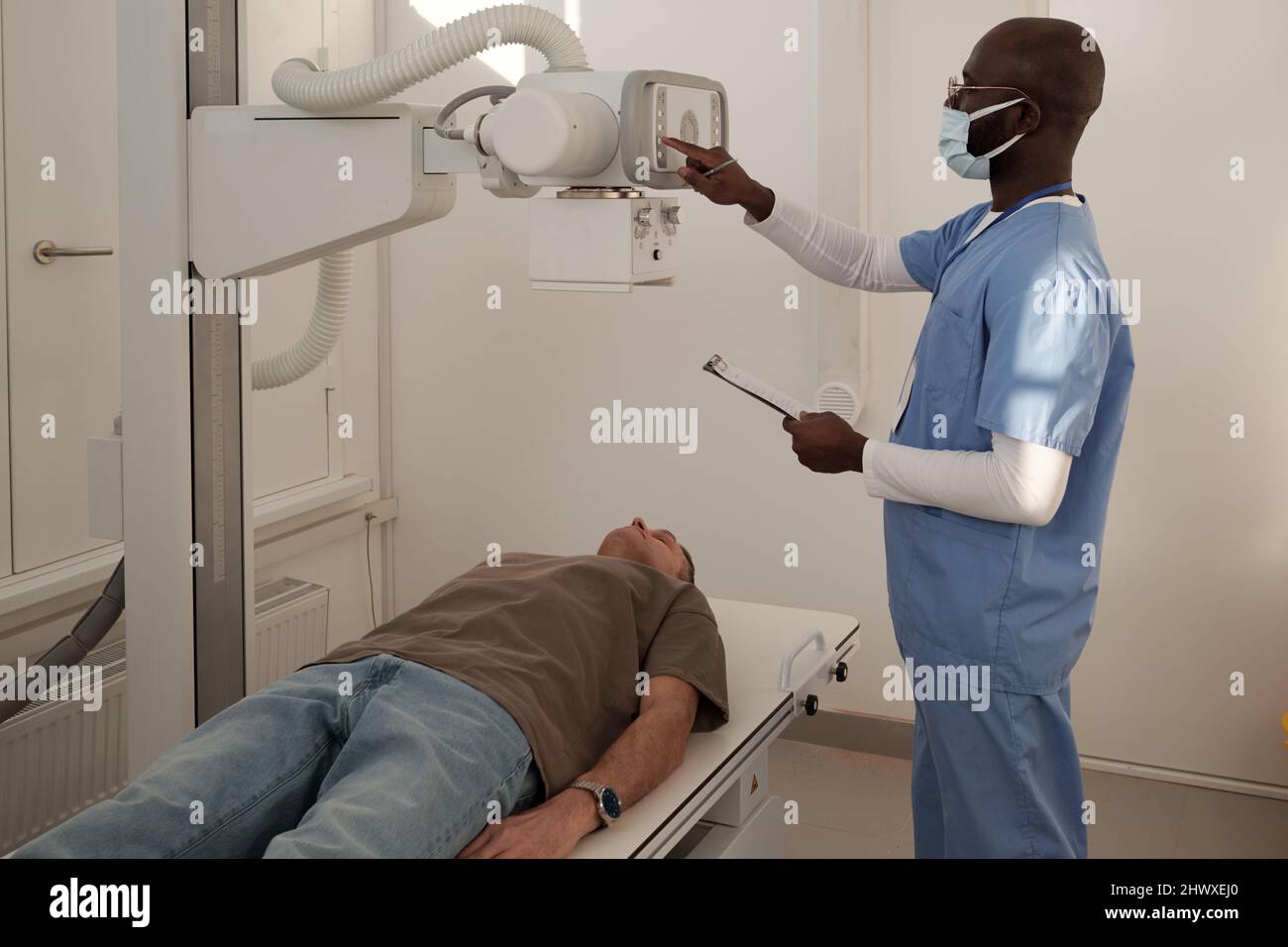 Jeune clinicien en uniforme et masque de protection allumer la machine à rayons X avant d'examiner un patient mâle mature à l'hôpital Banque D'Images