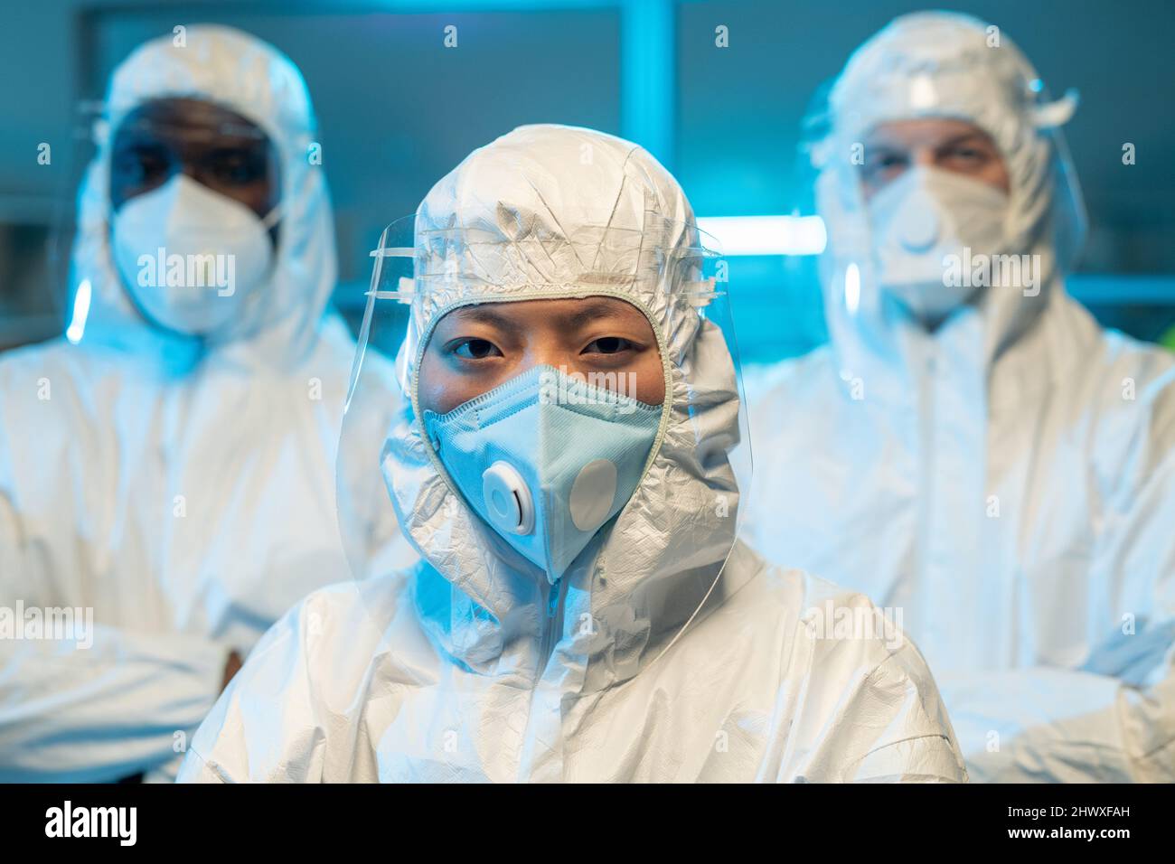 Trois scientifiques interraciaux dans des combinaisons de protection, des respirateurs et des écrans debout devant la caméra contre le laboratoire chimique Banque D'Images