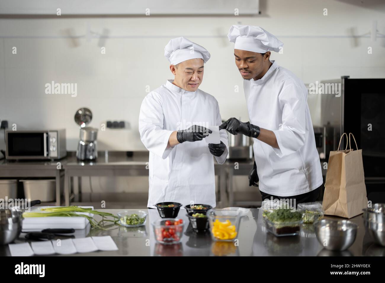 Les chefs asiatiques et latins lisent la commande imprimée tout en cuisinant des repas dans une cuisine professionnelle. Deux chefs de différentes nationalités cuisinent ensemble dans un restaurant Banque D'Images