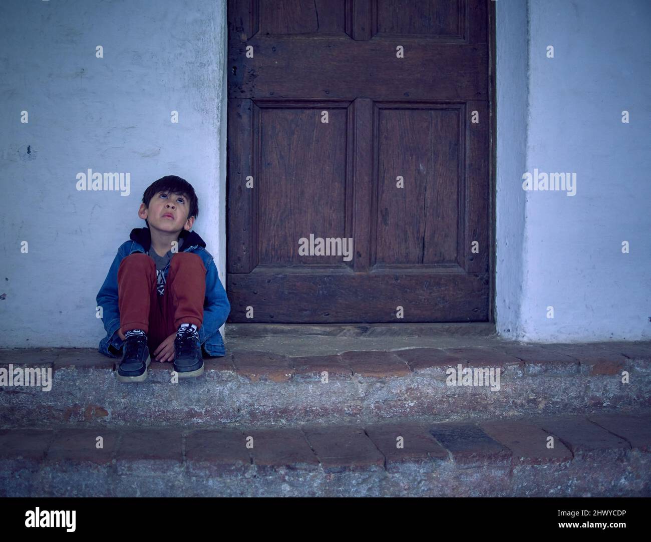 latino petit garçon est assis sur les escaliers près de la porte de la maison donnant vers le haut, attendant pensive et triste. Environnement horizontal et spectaculaire. Banque D'Images