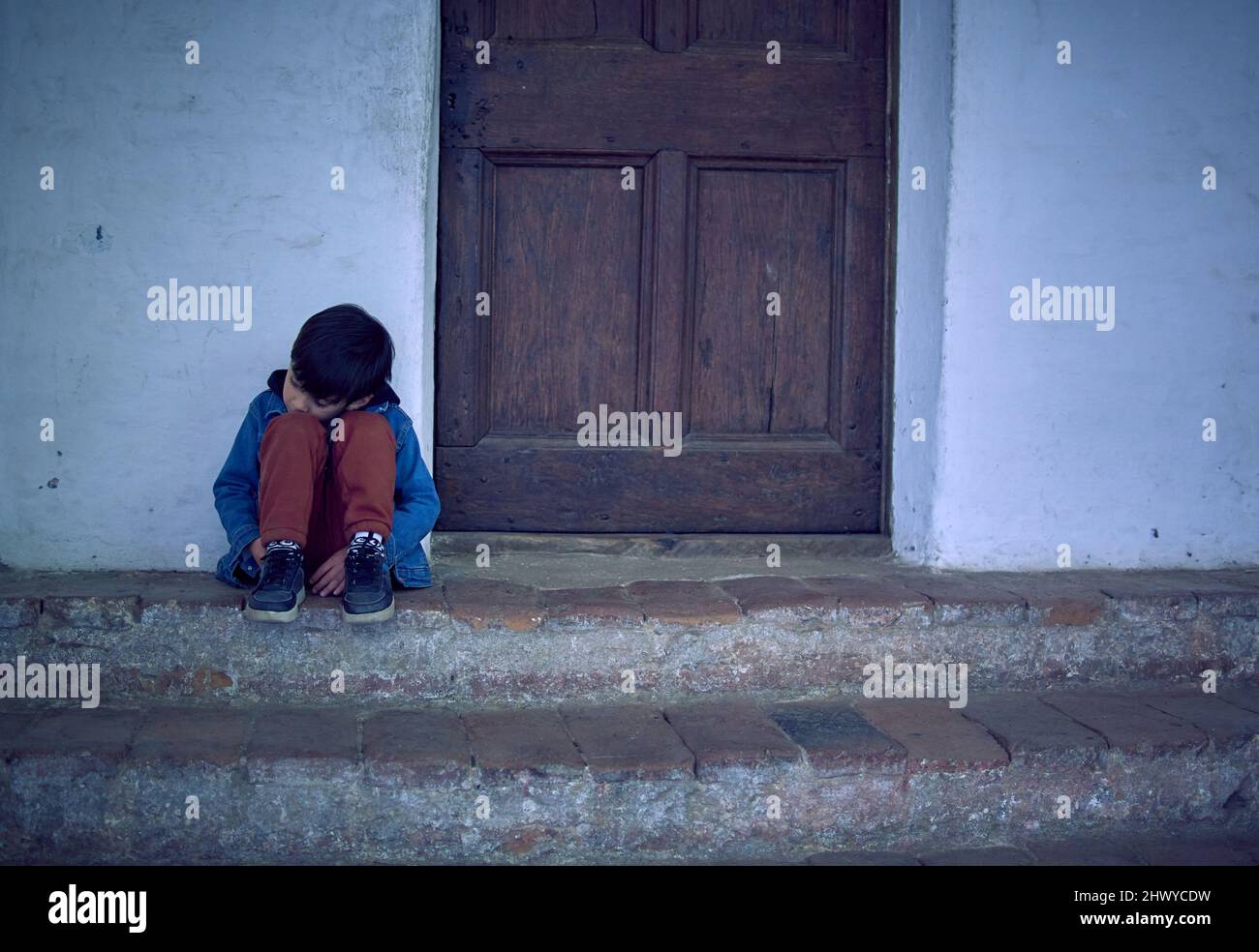 Melancholy Mixed Race Boy assis à l'extérieur de sa maison en colère et triste avec sa tête reposant sur ses jambes. Horizontal, latino enfant regardant vers le bas Banque D'Images