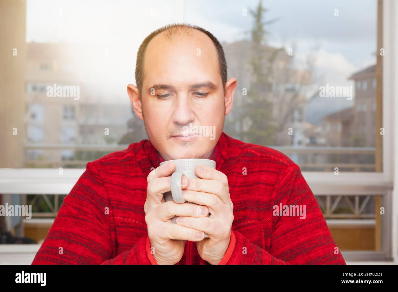Un homme à tête blanche dans un chandail rouge tient une tasse de café chaud, à la vapeur dans les deux mains, prêt à boire. En arrière-plan est une fenêtre et vous pouvez voir la construction Banque D'Images