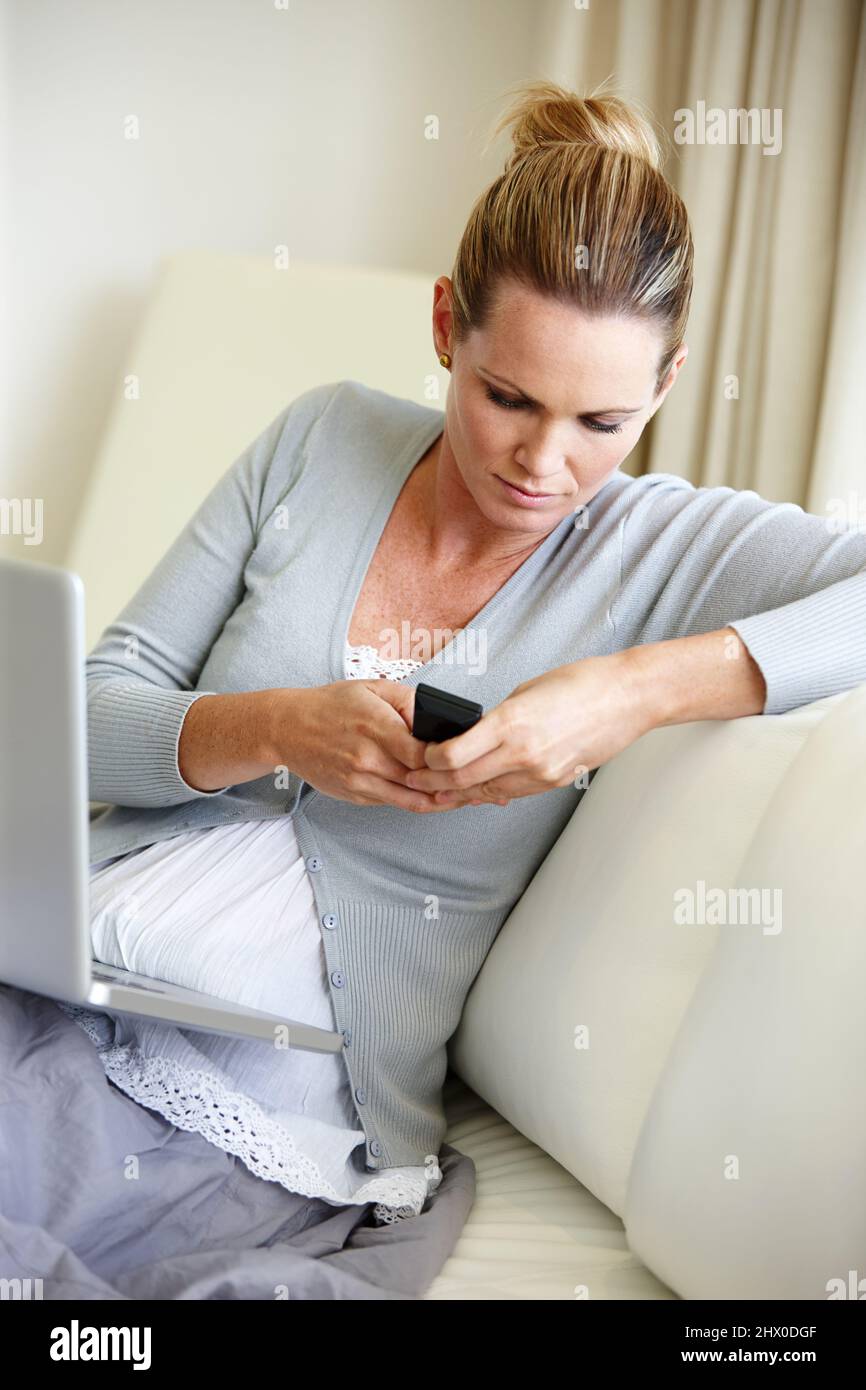 Merci pour la technologie. Une jeune femme se concentrant sur son téléphone portable tout en étant assise avec son ordinateur portable. Banque D'Images