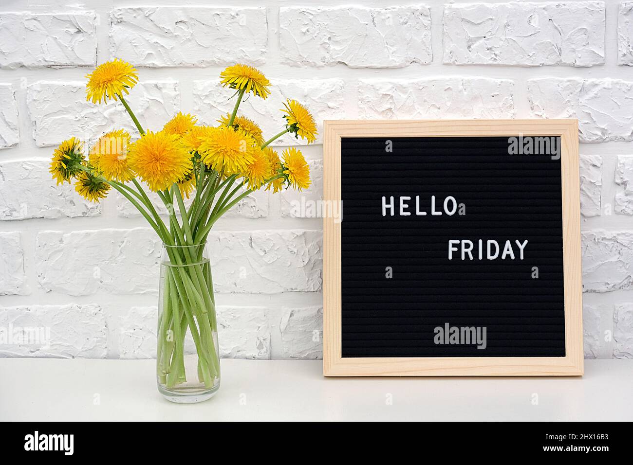Bonjour vendredi mots sur le tableau noir de lettres et le bouquet de fleurs de pissenlits jaunes sur la table contre le mur de brique blanche. Concept Happy Monday. Modèle pour Banque D'Images
