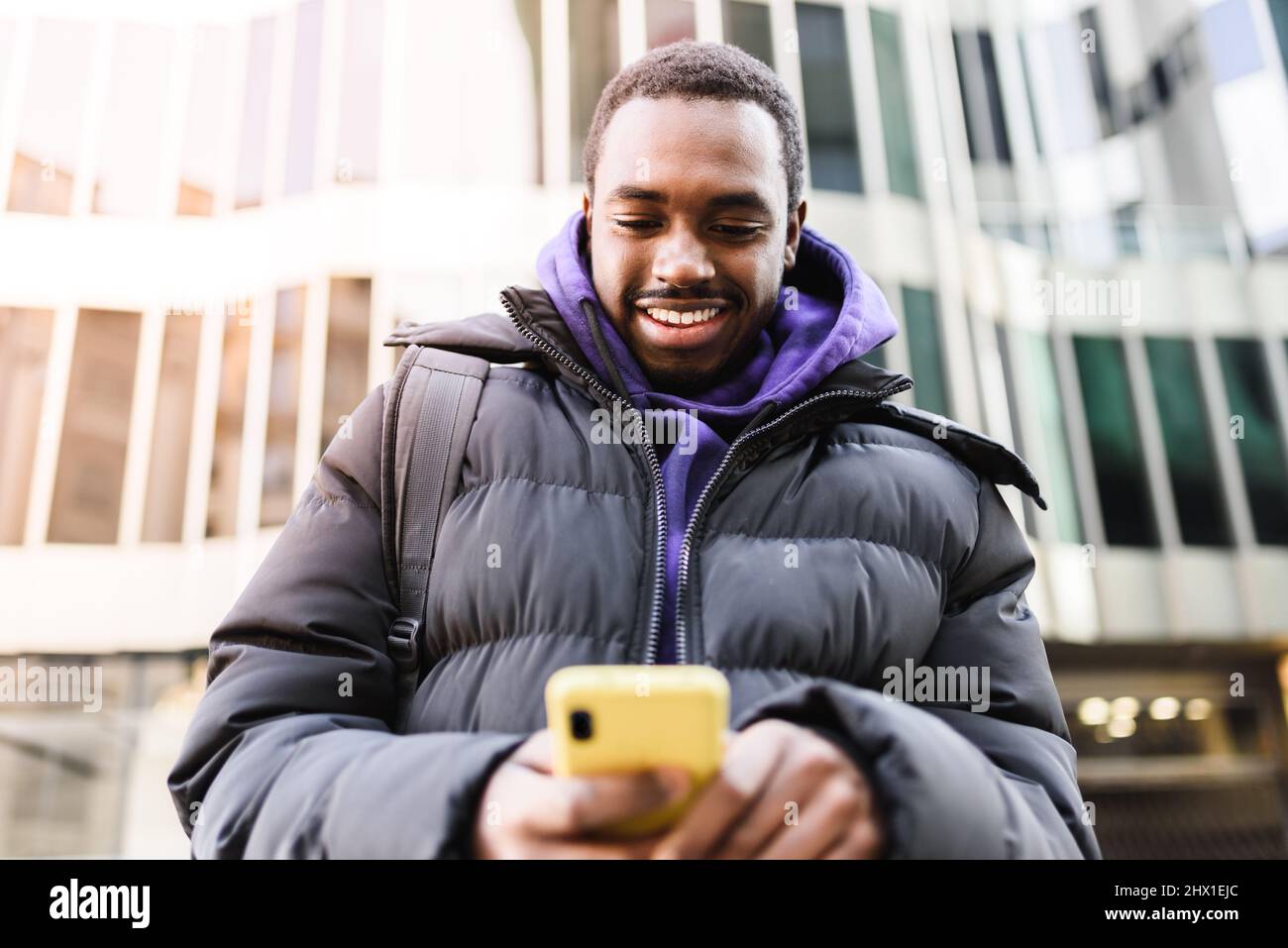 D'en-dessous, homme afro-américain joyeux en vêtements d'extérieur lisant et envoyant des messages texte sur smartphone tout en se tenant dans la rue moderne de la ville Banque D'Images