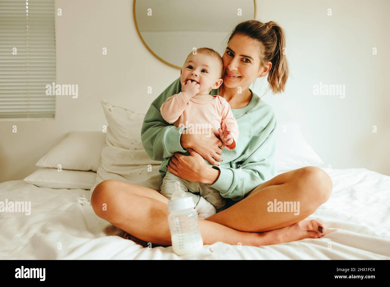 Bonne mère souriant à l'appareil photo tout en tenant son adorable bébé. Jeune mère gaie assise sur le lit avec son nouveau-né. Maman qui s'occupe créati Banque D'Images