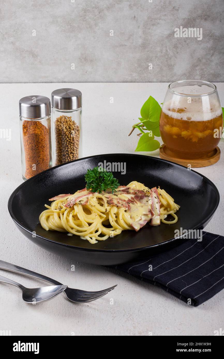 un spaghetti de pâtes cuit carbonara avec une sauce à la crème, une tranche de boeuf, de l'huile d'olive et du persil vert frais dans une assiette ovale noire avec une paire de pâtisseries Banque D'Images