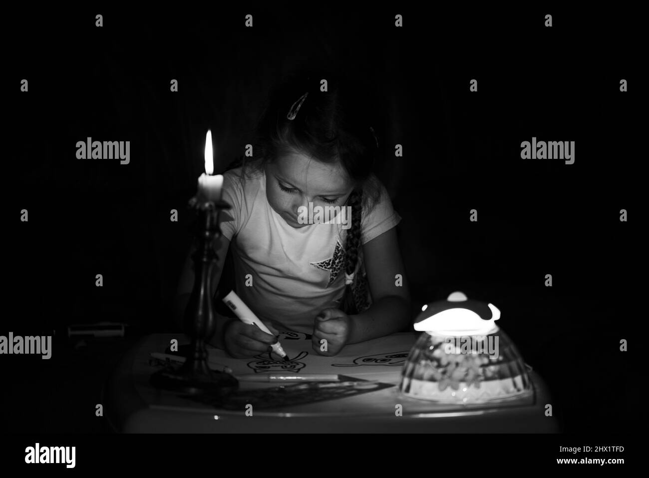 Une petite fille dessine des personnages de fantaisie près d'une bougie dans une pièce sombre. Enfant faisant ce qu'il préfère lors d'une panne de courant. En noir et blanc. Banque D'Images