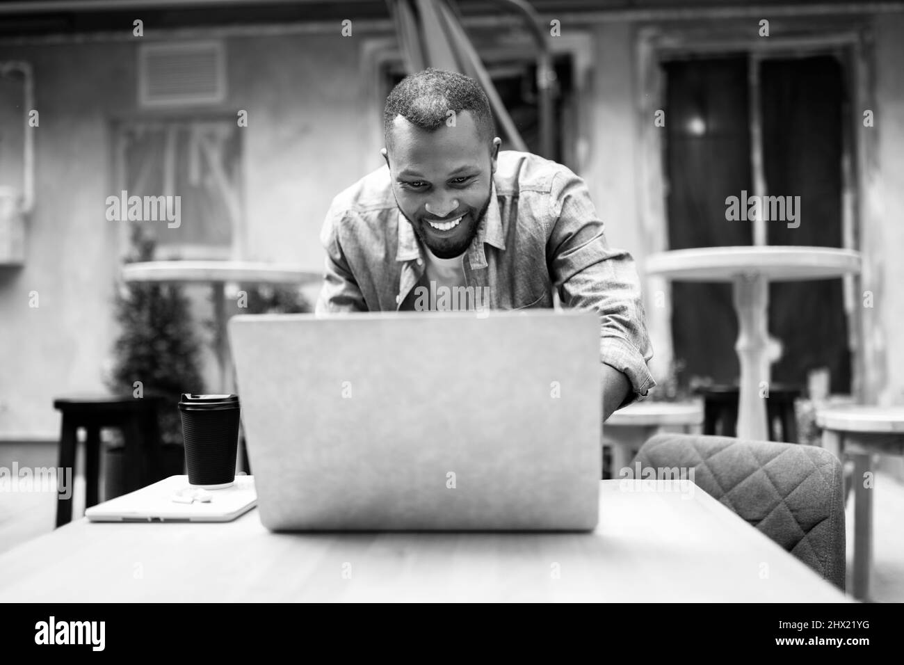 Homme d'affaires afro-américain pensif en vêtements élégants parlant sur téléphone mobile et en utilisant un ordinateur portable en plein air café, boire un café à emporter discuter avec humour Banque D'Images