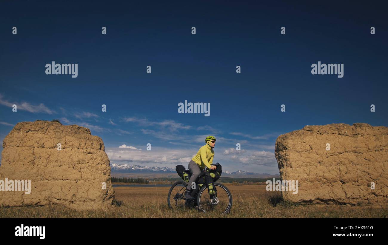 L'homme voyage sur des terrains mixtes en vélo avec bikepacking. Le voyage du voyageur avec des sacs de vélo. Sport bikepacking, vélo, vêtements de sport dans les couleurs vert noir. Montagne enneigée, arche en pierre. Banque D'Images