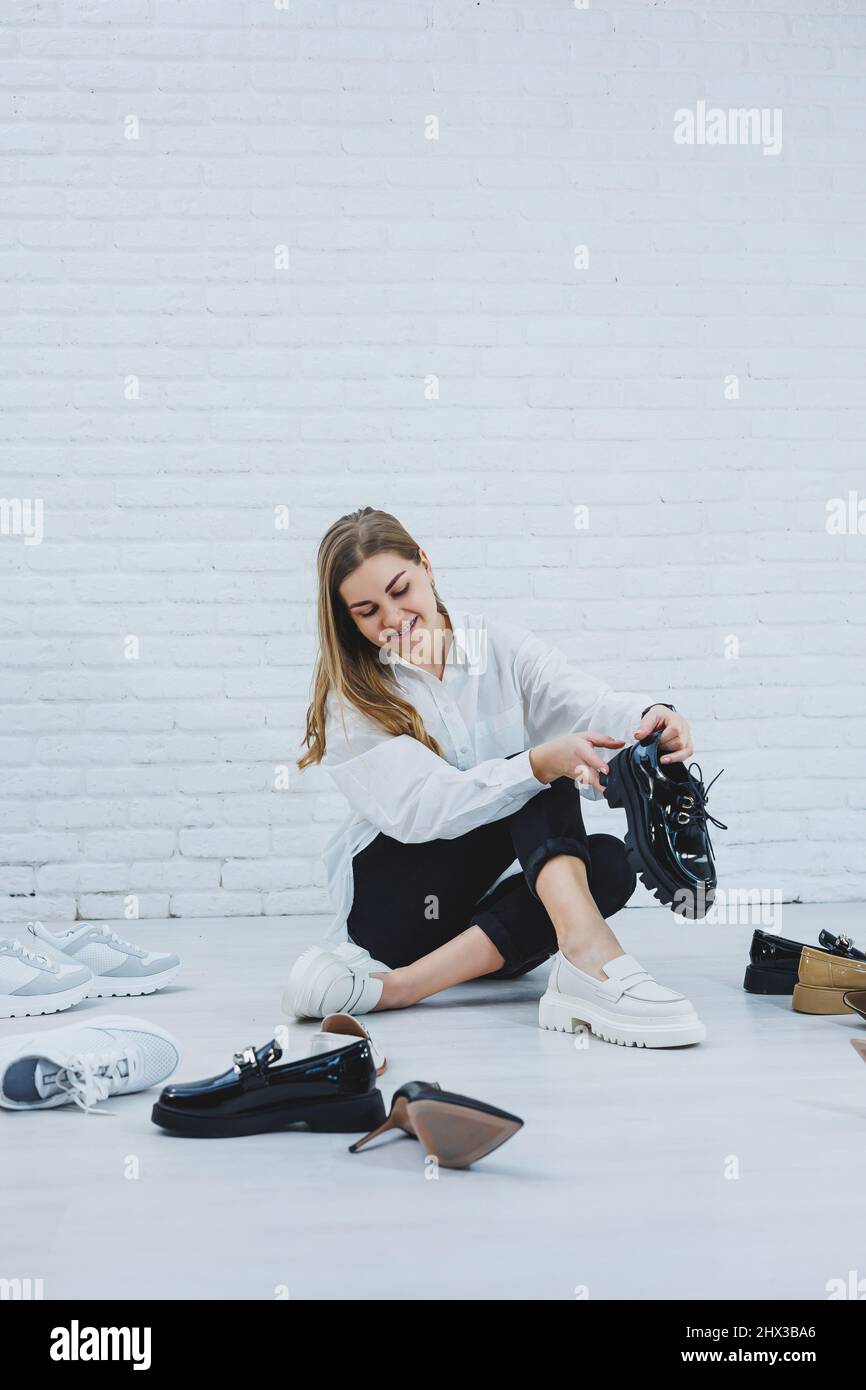 Une jeune femme s'assoit sur le sol parmi des chaussures et choisit de  nouvelles chaussures pour elle-même. Lady dans une chemise blanche et un pantalon  noir. Femme à chaussures blanches Photo Stock -