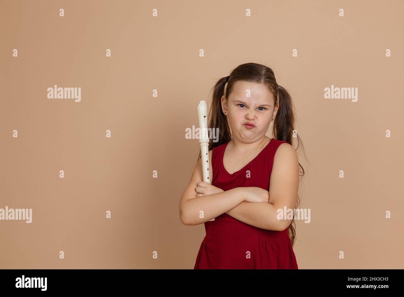 Fille en robe rouge avec la grimace mécontent tenir flûte avec les bras croisés, fond beige. Apprendre à jouer de l'instrument de musique de vent de bois. Banque D'Images