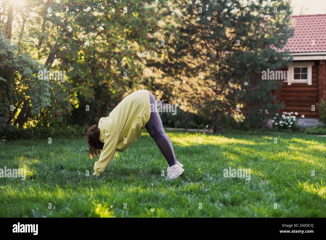 Obèse femme caucasienne s'exerçant à l'extérieur dans des vêtements de sport avec les pieds et les mains mis sur terre sur l'herbe fraîche sur l'arrière-cour avec maison de campagne en bois et de haut Banque D'Images