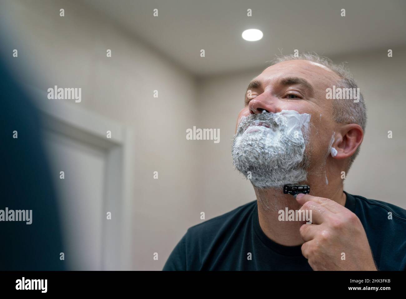 Homme mûr aux cheveux gris recoiffant lui-même sa barbe à la maison à l'aide d'un rasoir Banque D'Images