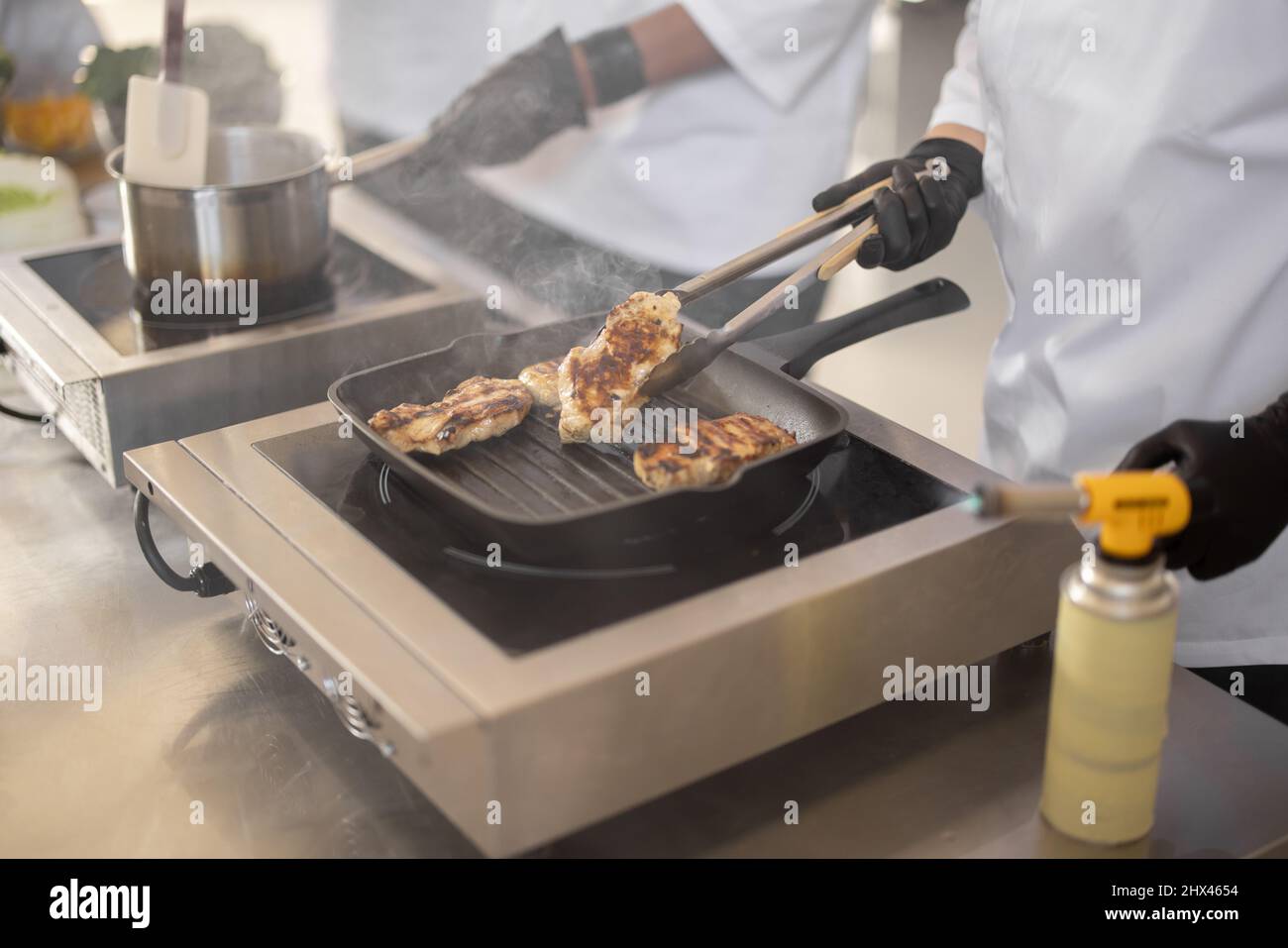 Cuisiniers en gants et uniforme préparant les aliments dans une cuisine professionnelle, friture de la viande et de la sauce sur les tables électriques, vue rapprochée du processus de cuisson Banque D'Images