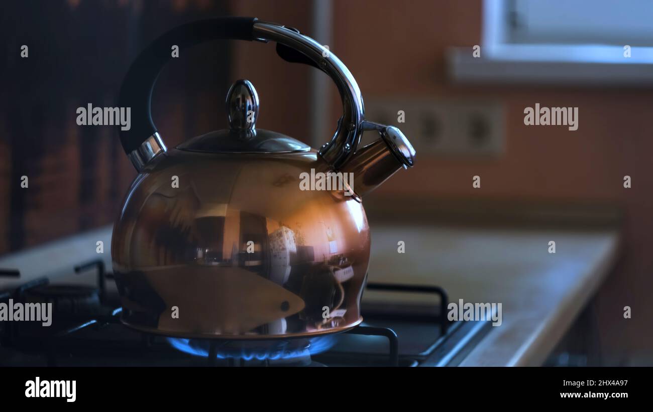 Gros plan d'une bouilloire à thé en acier inoxydable sur une cuisinière à gaz flamboyante. Bouilloire de chauffage avec le reflet de la cuisine sur sa surface métallique, préparation o Banque D'Images