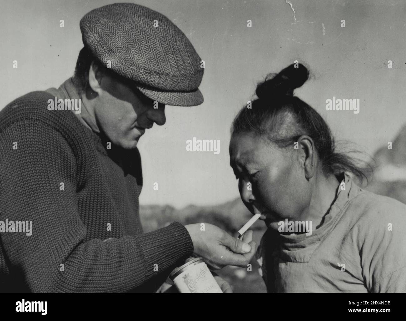 Dans le Nord gelé avec un Arctic Commuter -- une femme esquimau âgée dans l'un des rares établissements de l'est du Groenland accepte avec gratitude une cigarette « sur mesure » et une lumière de l'un des membres de l'équipage de Caption Bob Bartlett. Le bouton de cheveux ressemble au shimadi japonais. 17 octobre 1939. (Photo par Wide World photos). Banque D'Images