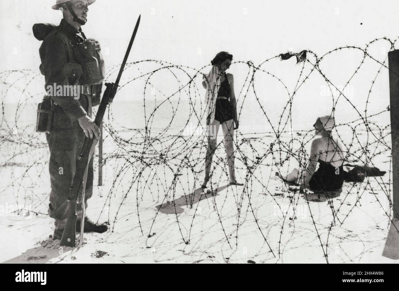 Sur Une plage britannique : deux Vacationistes se détendent sur une plage non identifiée en Angleterre, des sources britanniques se sont assis pour décrire cette image, comme un Tommy, les yeux tout droit devant, se tient à la garde d'une barrière construite comme une défense contre une éventuelle invasion de la côte française. 21 septembre 1940. (Photo par photo de presse associée). Banque D'Images