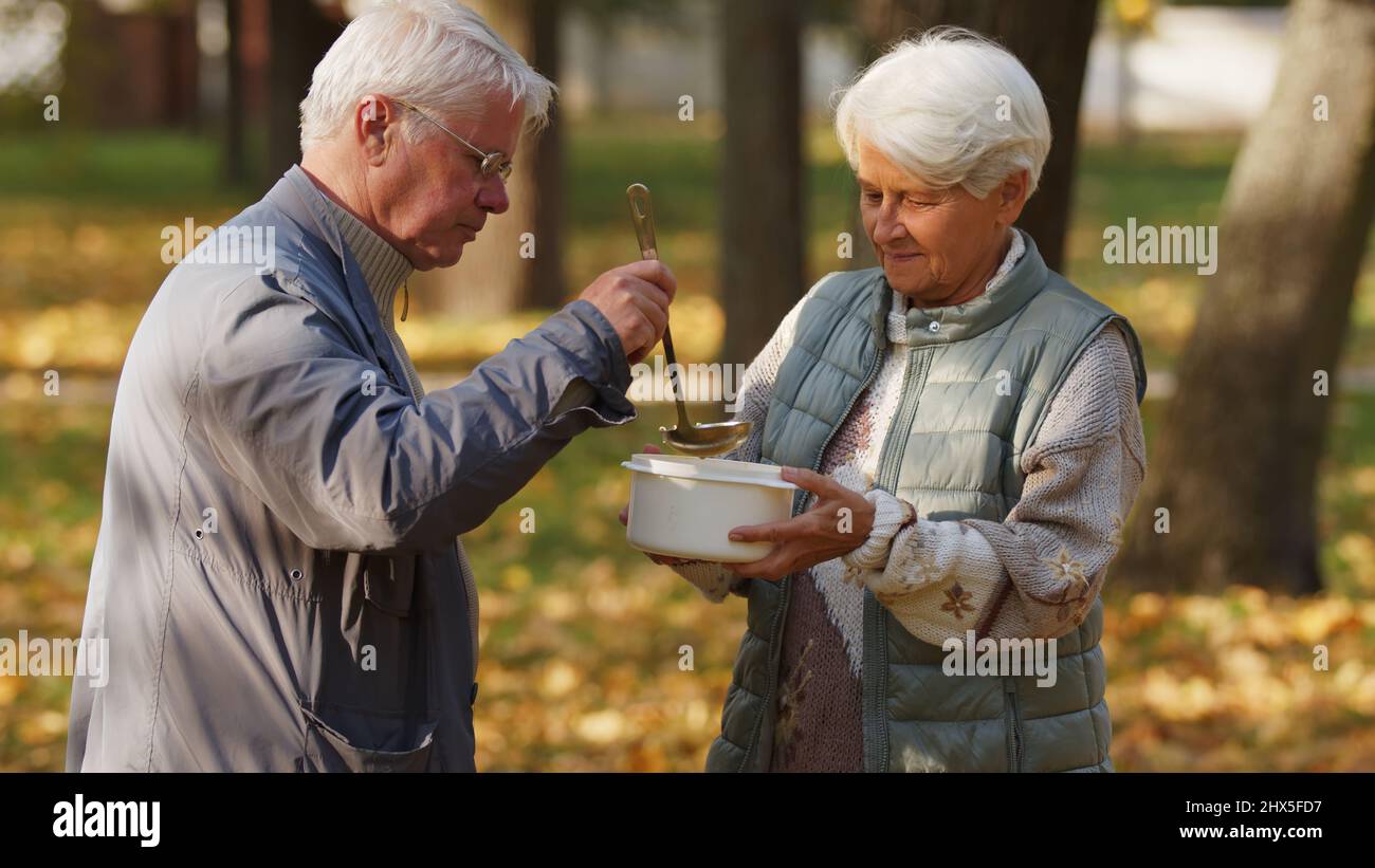 Généreux homme caucasien âgé à poil gris bénévoles dans une organisation caritative et verse de la soupe chaude dans le bol blanc de la femme sans abri âgée. Photo de haute qualité Banque D'Images