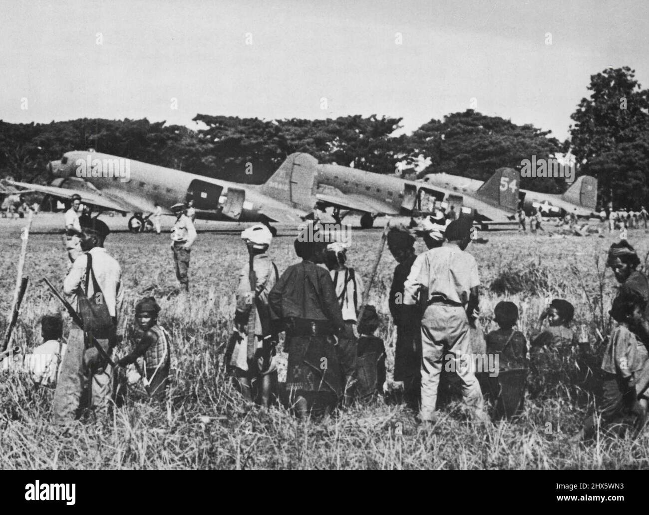 Surveillance birmane déchargement des fournitures de combat des avions de cargaison des États-Unis -- Les résidents birmans qui étaient sortis des jungles à volant pour observer les précédents, se tiennent au bord de cet aéroport récemment achevé dans le nord de la Birmanie et regardent curieusement pendant que les avions de cargaison de l'armée de l'air américaine de 10th sont déchargés. Les grands transports avaient atterri plus tôt avec des fournitures pour les forces alliées roulant contre les Japonais dans la région. L'armée de l'air de 10th a non seulement apporté des fournitures, mais a également apporté un soutien tactique aux Chinois Banque D'Images