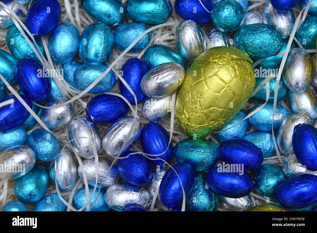 Pile de feuilles pastel colorées, enveloppée de chocolat, œufs de pâques en bleu, argent et turquoise avec un gros œuf jaune au milieu, sur fond pâle. Banque D'Images