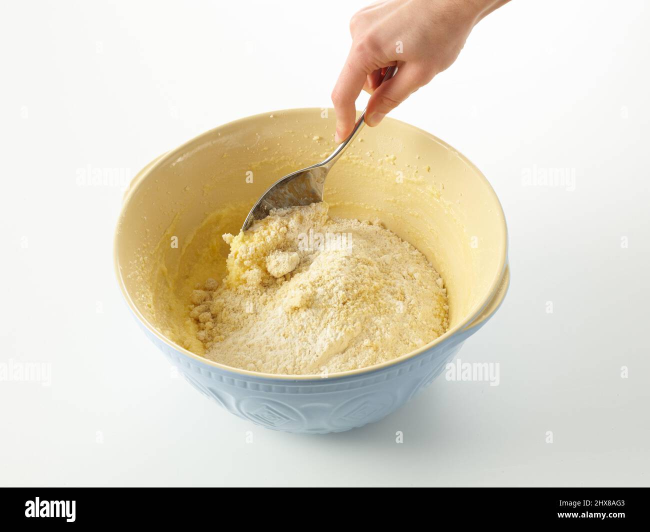 Incorporer la farine, la poudre à pâte et les amandes moulues dans un mélange d'œufs, de sucre en poudre, de beurre et d'eau de rose, afin de faire un gâteau peint aux roses et aux amandes. Utiliser une grande cuillère en métal dans un bol à mélanger bleu. Banque D'Images
