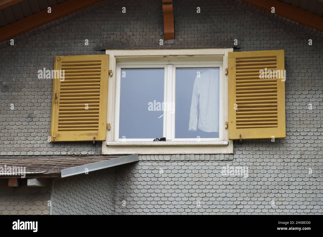 Fenêtre avec volets ouverts. Une chemise blanche est suspendue dans la fenêtre. Façade de maison en Suisse. La façade est recouverte de bardeaux en bois. Banque D'Images