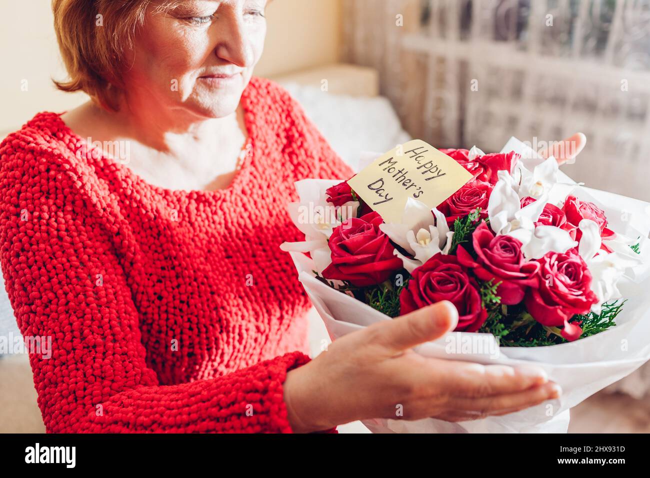 Bonne surprise de la fête des mères. La femme âgée tient un bouquet de roses rouges et des fleurs d'orchidées à la maison avec une carte de vœux. Cadeau pour les vacances de printemps Banque D'Images