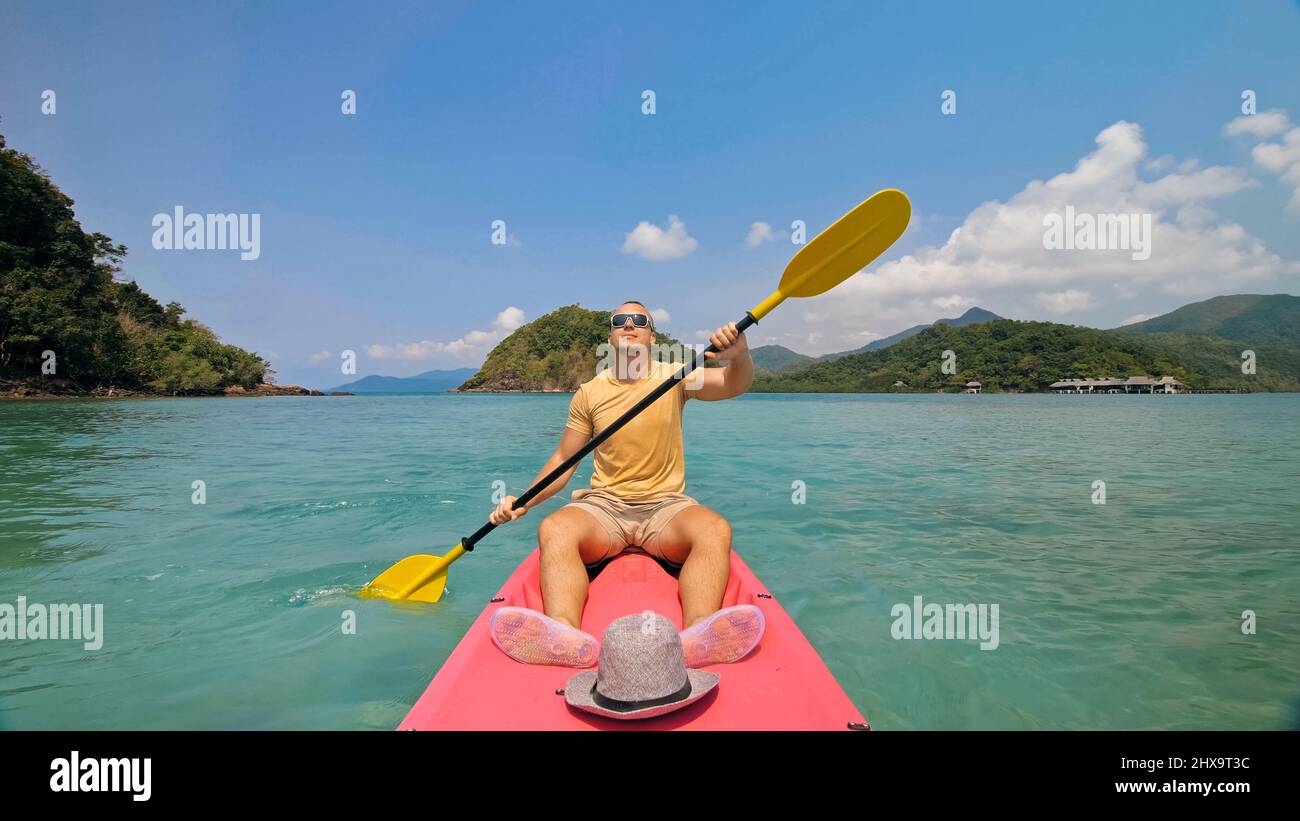 Jeune homme avec des lunettes de soleil et un chapeau rows en plastique rose canoë le long de la mer contre des îles vertes vallonnées avec des jungles sauvages. Voyager dans les pays tropicaux. St Banque D'Images