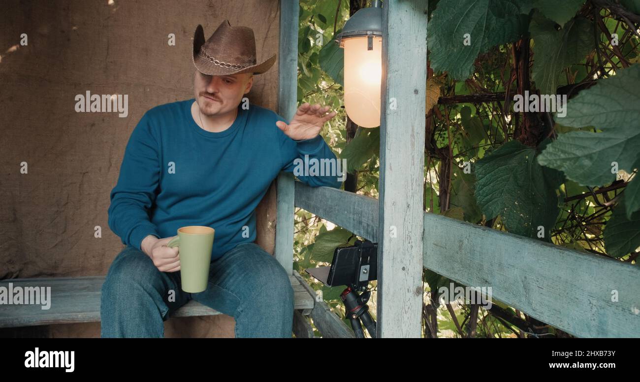 Un cowboy sur le porche boit le café d'une tasse et communique en ligne par téléphone Banque D'Images