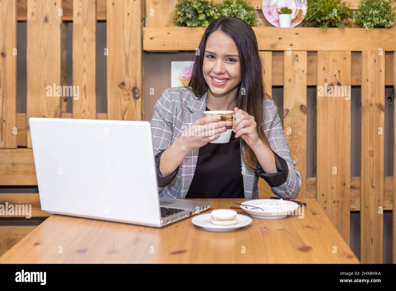 Femme indépendante et optimiste, blouson à carreaux, buvant un café chaud tout en étant assise à table avec un netbook et un macaron sur l'assiette et en regardant l'appareil photo Banque D'Images