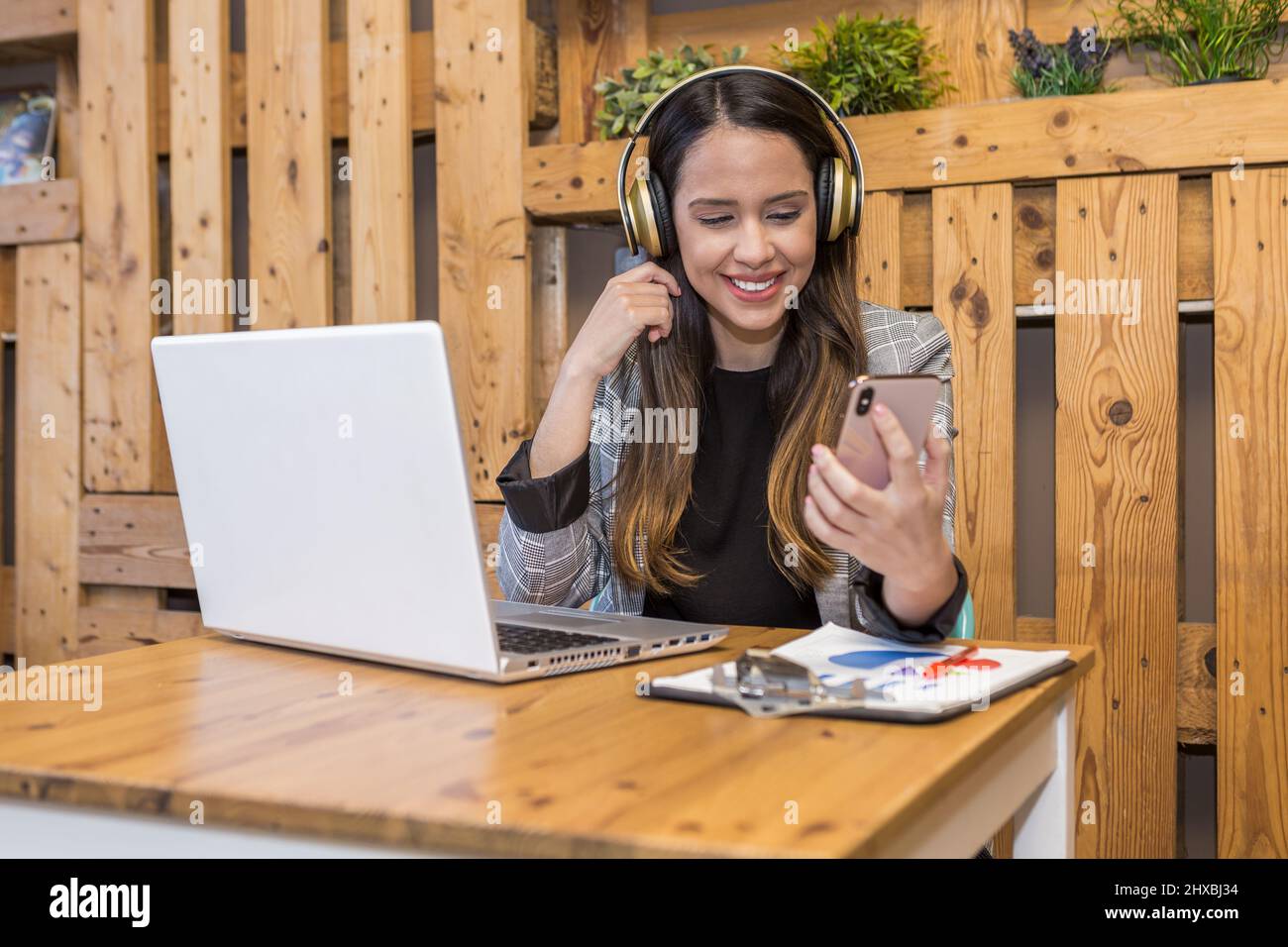 Une employée à distance positive dans un casque sans fil assis à une table en bois et vérifiant les messages sur un téléphone portable pendant le travail en ligne sur un netbook Banque D'Images