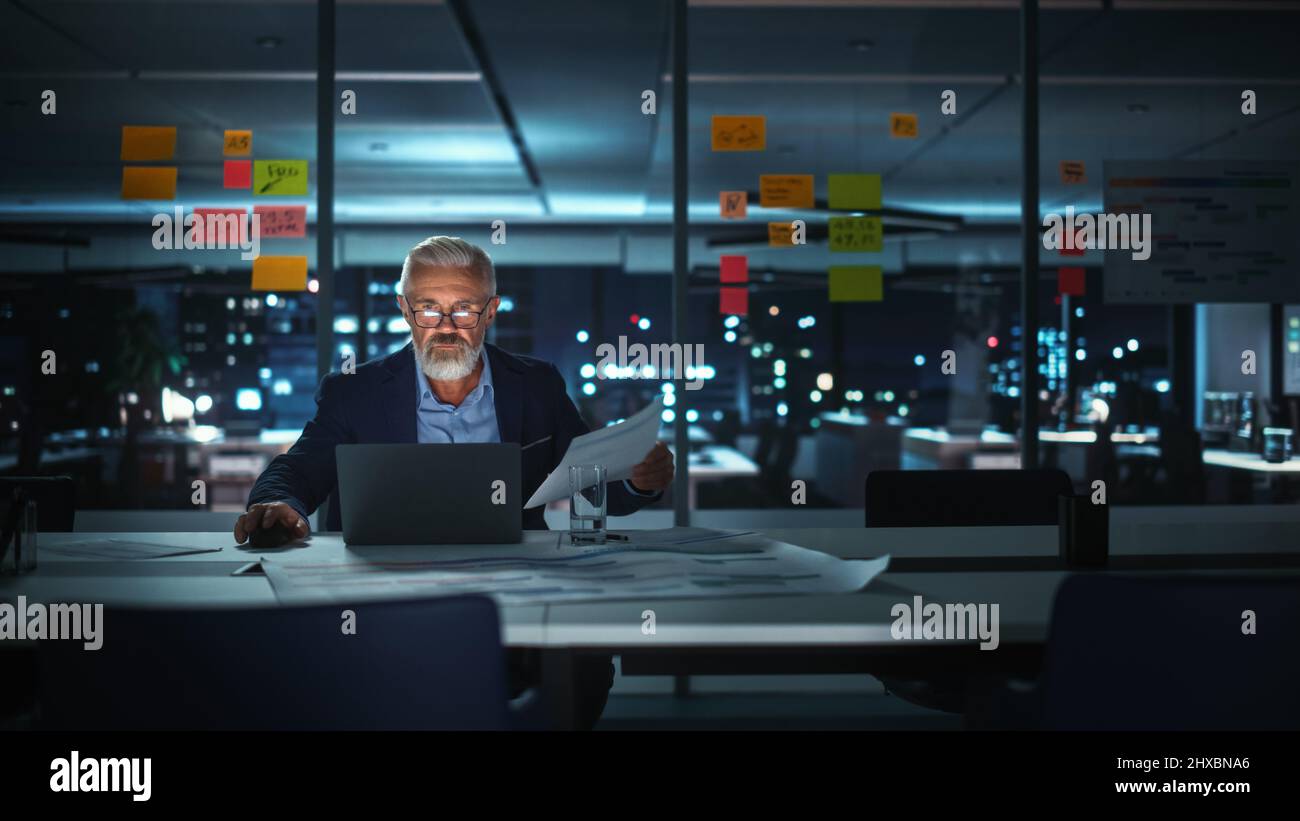 Un homme d'affaires senior prospère travaillant sur un ordinateur portable au bureau de Big City tard dans la soirée. Analyste de placement contrôle de gestion financière repo Banque D'Images