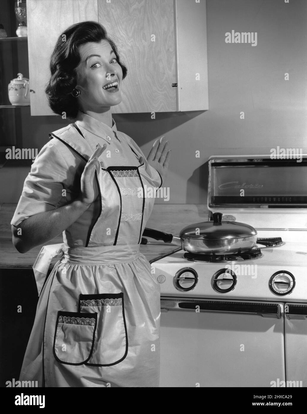 Femme à son milieu des années 30, bien habillée avec un tablier de cuisine debout devant son four avec les mains vers le haut et les pouces sous son tablier en souriant Banque D'Images