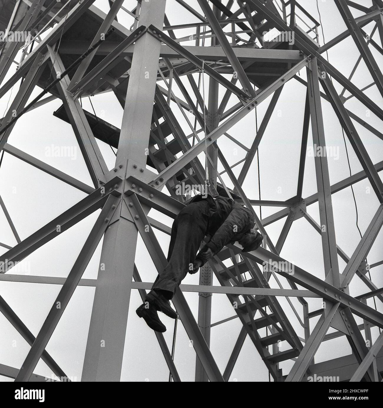 1950s, historique, un steelworker sur un palan étant descendu d'une tour d'acier ou d'un pylône, au complexe d'acier Abbey Works, Port Talbot, pays de Galles, Royaume-Uni, dans un exercice de santé et de sécurité. Banque D'Images