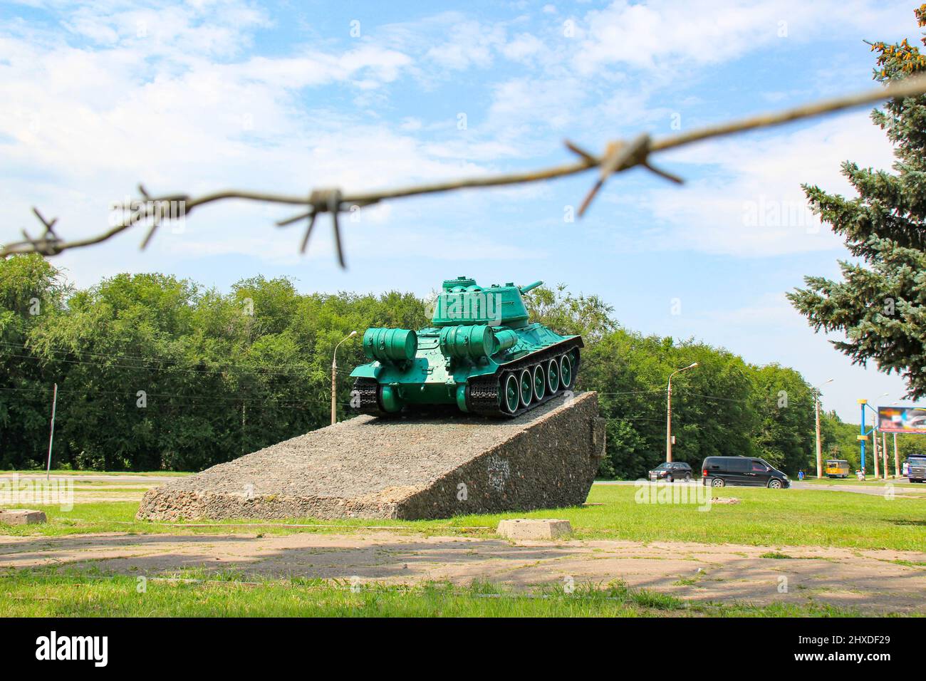 Monument à l'ancien réservoir soviétique T-34. Un char militaire se dresse sur un piédestal sur fond de ciel bleu, de barbelés, d'arbres verts, de rue de la ville. Banque D'Images