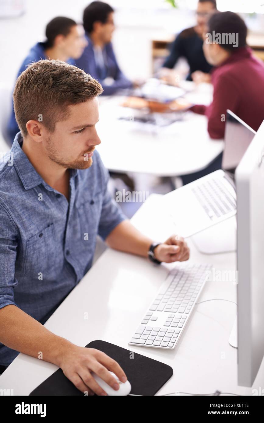 Les entreprises modernes sont innovantes et en ligne. Photo d'un jeune designer travaillant sur son ordinateur avec des collègues en arrière-plan. Banque D'Images