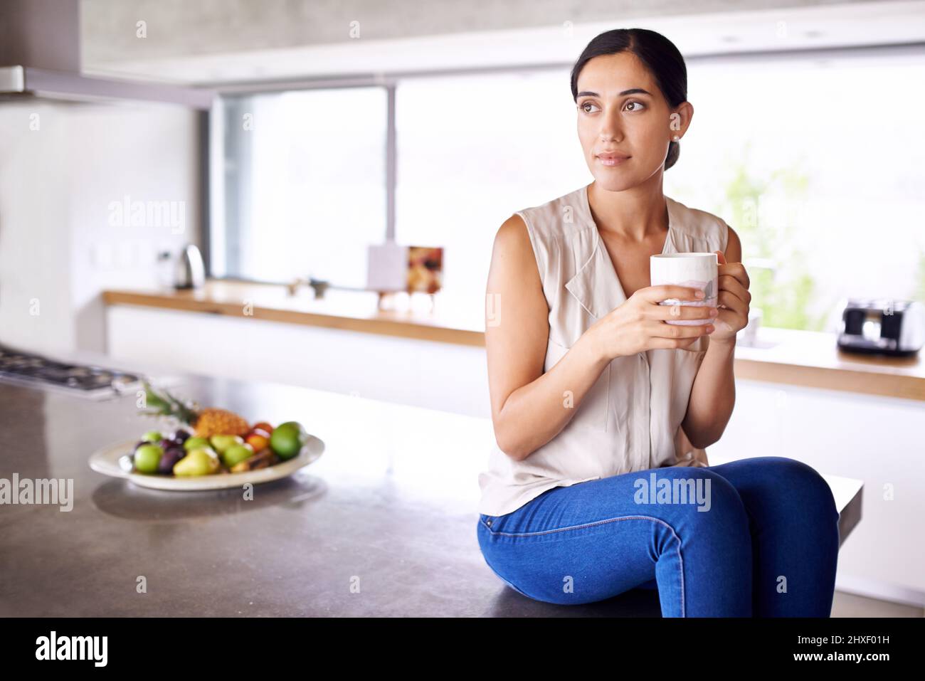 Savourez une tasse de café chaud. Photo d'une jeune femme attrayante assise sur le comptoir de sa cuisine et buvant du café. Banque D'Images