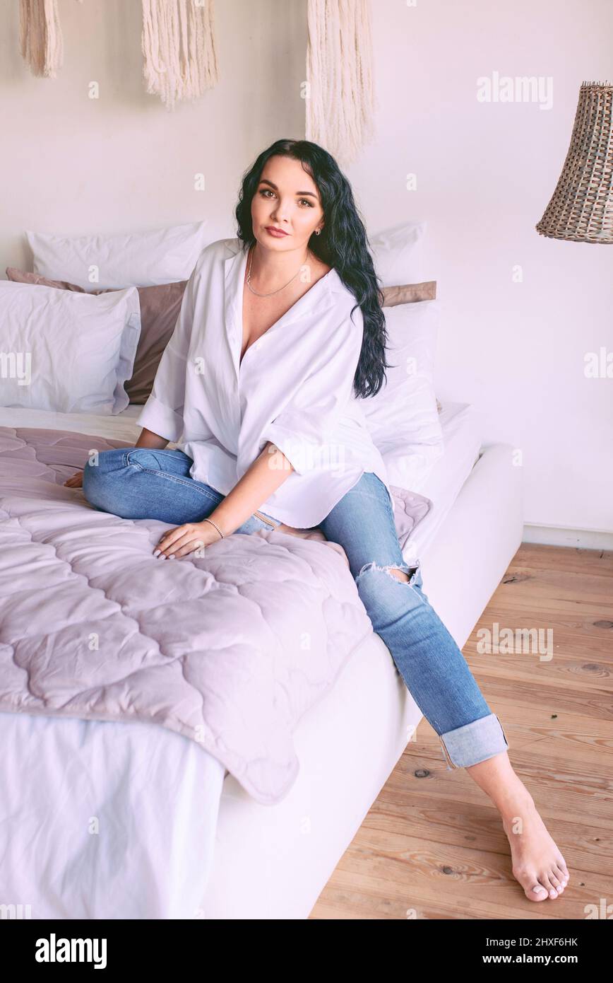 Portrait de belle femme à cheveux foncés en chemise blanche et jeans assis triste sur son lit. Émotions, crise, maison, concept intérieur Banque D'Images