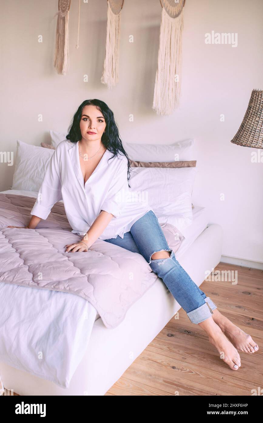 Portrait de belle femme à cheveux foncés en chemise blanche et jeans assis triste sur son lit. Émotions, crise, maison, concept intérieur Banque D'Images