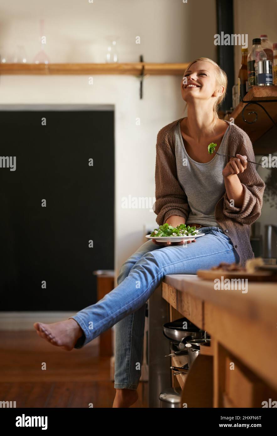 Shes aimant son nouveau régime. Photo d'une jeune femme attirante assise sur son comptoir de cuisine en riant tout en mangeant une salade. Banque D'Images